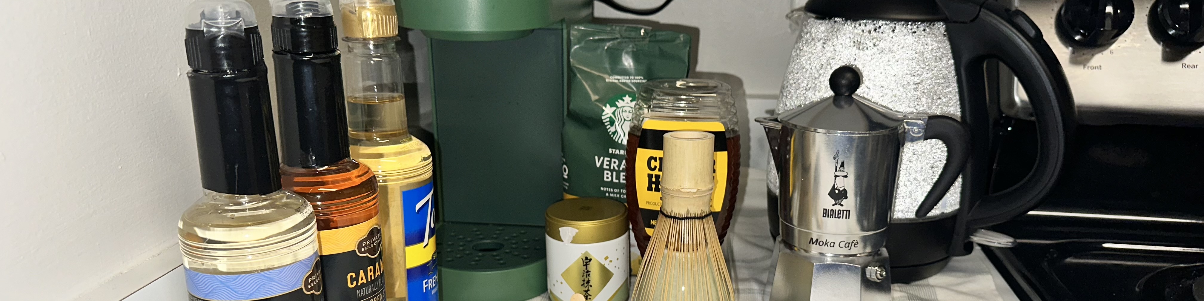 Several items including a coffee machine, a kettle, a bag of coffee, a moka bot, syrups, and a can of matcha powder.